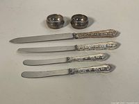 Four knives with ornate silver handles and two silver napkin rings arranged flat on a light surface