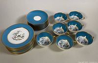 Set of 10 dessert plates stacked with 7 teacups and 8 saucers arranged beside them showing blue, gold trim, and peacock floral center designs.