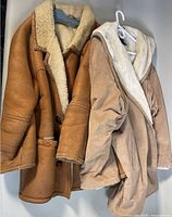 Two sheepskin and leather coats shown hanging on hangers, one leather with buttons and one suede with hooded sheepskin lining showing.
