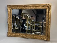 Full rectangular mirror showing gold ornate floral Baroque frame standing against a neutral background, reflecting room shelving and items.