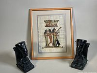 Pair of black Queen Nefertiti chalkware bust bookends flanking a framed Egyptian papyrus artwork of three musicians.
