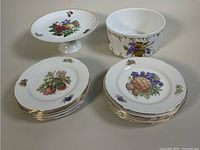The full set showing twelve fruit plates, one pedestal cake plate, and one decorative bowl, all porcelain with fruit and grape designs and gold trim.