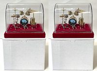 Front view of two miniature drum set models inside clear plastic dome display cases with red bases, placed on white boxes.