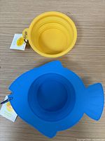 Top-down view of yellow round collapsible pet bowl and blue fish-shaped collapsible pet bowl, showing their colors, shapes, tags and clips.