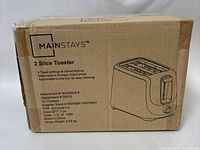 Photo of the original cardboard packaging box of a Mainstays 2 slice toaster showing product branding, basic features including 6 toast settings, cancel feature, wide slots, and removable crumb tray.