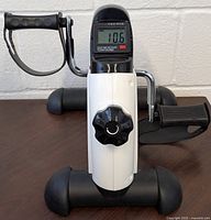 Front side view showing adjustable resistance knob, LCD display with exercise metrics, and foot pedal with strap.