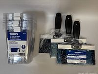 Set of 5 packs of clear plastic pail liners and 3 blue carbon steel taping knives with black handles shown stacked together