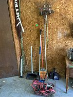 Photo of brooms, rakes, dustpans, badminton net rolled up, and cables hanging or on floor against wood wall.