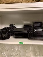 Shelf displaying multiple black ceramic tea cups stacked with matching saucers alongside rectangular plates and square bowls in mixed styles.