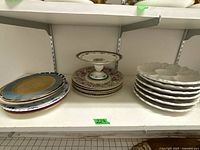 Multiple china plates, pedestal serving plate, and white bowls displayed on a shelf.
