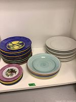 Stack of multicolored handmade Italian plates, white plates, and floral side plates in a corner on a white shelf.
