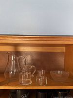 Shelf with a large clear pitcher with handle and spout, smaller glass pitcher, small cylindrical glass container, and a medium clear glass bowl.