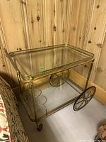 Front side view of a gold-tone metal bar cart showing glass top shelf and lower shelf with circular holders, large spoked wheels on one side, and smaller wheels on the other.