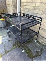 Full view of black iron bar cart showing decorative edge detail, two mesh shelves, and different wheel sizes.