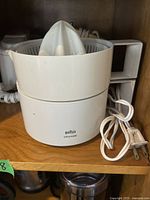 White Braun electric citrus juicer with power cord coiled in front on wooden shelf