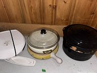 Shot showing white George Foreman grill, ceramic pot with glass lid, and two large black enamel-coated cast iron pots, one with heavy wear.