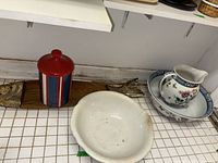 View of large off-white ceramic serving bowl, red and blue striped tin container and wooden fish shaped board placed on tiled counter.