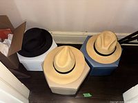 Three men's hats placed on top of three hat boxes against a wall; two are natural straw fedoras with black bands, and the third is a black fedora.