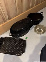Photo showing four pieces: black speckled oval roasting pan with lid, black round cast iron pot with lid, black roasting tray with spikes, and metallic bundt cake pan.