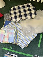 Various placemats arranged on a green carpet. Includes blue and white checkerboard placemats with nautical symbols, striped placemats in pastel colors, and translucent floral patterned placemats.