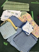 Assortment of placemats, kitchen towels, and pot holders displayed in a basket including white towels with dragonfly prints, blue towels, floral patterned tea towels, blue and white checked placemat, and light blue quilted pot holders.