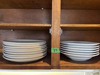 Two stacks of white plates on a wooden shelf, one stack contains 10 plates and the other 7 plates, showing overall good condition.