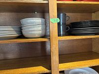 White ceramic plates and bowls stacked on left side, dark black and grey plates and two mugs stacked on right side on wooden shelf.