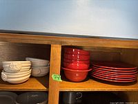 Wooden cabinet shelf with white ceramic bowls on the left and red ceramic plates and bowls on the right.
