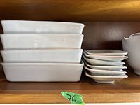 Three stacked rectangular white ceramic serving trays and a stack of eight smaller white ceramic plates with slightly raised edges.