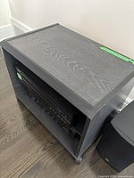 Angled view of small grey wood-grain table with open front shelf and visible wheels, holding some electronics inside.