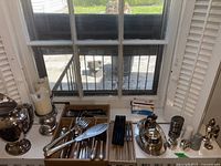 Overview of silver pieces on windowsill including pitcher, candlesticks, cutlery tray, dome, and shakers