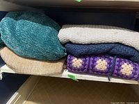 Stack of five throw blankets showing various textures and colors including turquoise, beige, blue, cream, and purple granny square design.