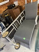 Two grey outdoor lounge chairs stacked and leaning against a wall in a garage. Both have grey mesh seats and backs, silver metal frames, and black/tan wheels at the foot end.