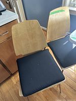 Two wooden chairs with light wood backs and black seat cushions shown side by side in a kitchen setting.