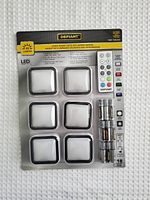 Front view of the blister pack showing six white square LED accent lights and a remote control. Batteries are included in the packaging.