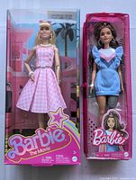 Front view of two unopened Barbie dolls in original packaging. One doll wears pink gingham dress and the other wears a blue dress.