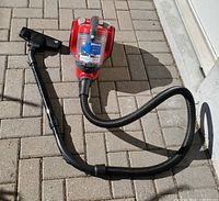 Full view of the red Dirt Devil Breeze vacuum cleaner with attached hose and separate floor cleaning nozzle on a paved surface.