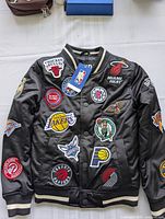 Front view of black NBA kids jacket with numerous embroidered NBA team logos including Chicago Bulls, Miami Heat, Los Angeles Lakers, Boston Celtics, and more on the front and sleeves.