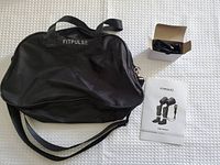 Black Fitpulse carry bag with shoulder strap, power adapter in open box, and instruction manual on white surface.
