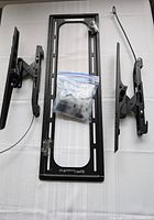 Top-down view of the Sanus TV mount components laid out on white fabric. Includes two adjustable mounting arms, rectangular mounting bracket back plate with a plastic bag of screws and hardware.