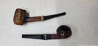 Two vintage tobacco pipes laying flat on a white surface, showing one rusticated bowl and one carved bowl, both with black stems.