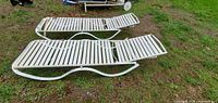 Two white metal slat chaise loungers placed on grass, showing side profile and adjustable backrest visible.