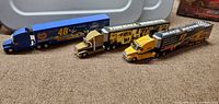 Three diecast NASCAR-themed tractor trailers lined up on carpet showing various sponsor branding and colors.