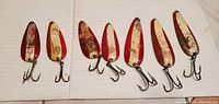 Seven red and white metal fishing spoon lures with treble hooks laid out in a row showing their fronts. The lures are approximately 4-5 inches long and show paint wear and aging.