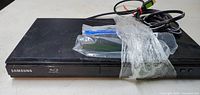Front view of black Samsung Blu-ray player with power cable and small accessories wrapped in plastic.