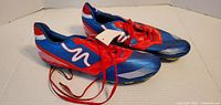 Pair of new blue and red soccer cleats shown from side angle with visible tags and red laces.