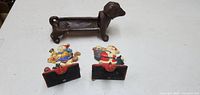 Photo showing two heavy metal Christmas stocking holders with festive holiday designs and a cast iron sausage dog figurine/doorstop on a white surface.
