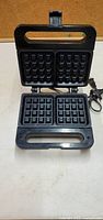 Front view of waffle maker showing black rectangular waffle plates and handle, power cord visible.