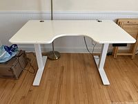 White L-shaped adjustable height desk with cable management hole and leg support on wooden floor.