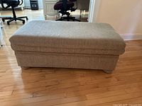 Front angled view of rectangular beige fabric ottoman on hardwood floor, showing fabric texture and cushioned top.